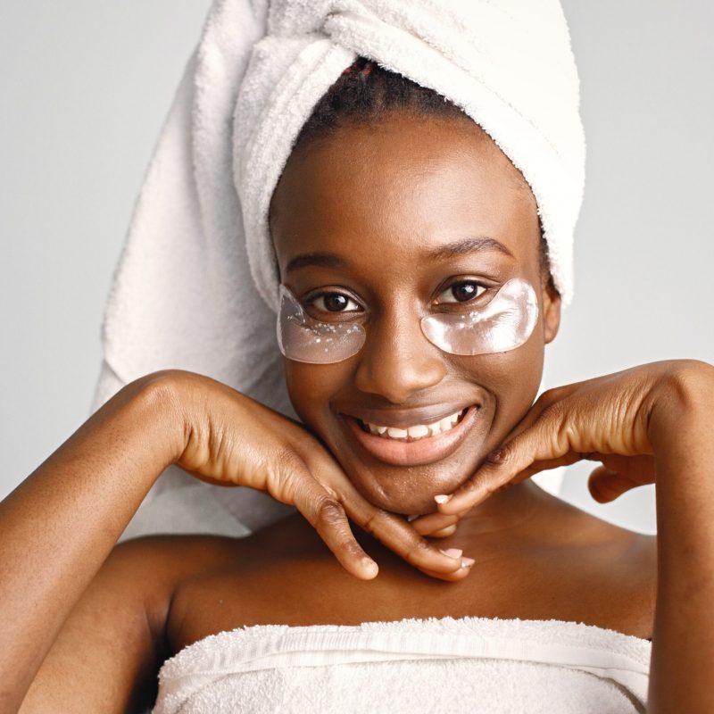 Black young woman standing isolated on white background with patches on her face. Girl has her hair wrapped in a towel. Black  woman enjoy after body treatment.