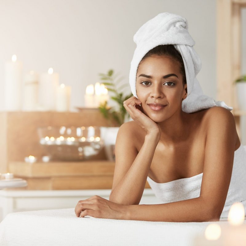 Portrait of beautiful african girl with towel on head smiling looking at camera resting in spa salon.